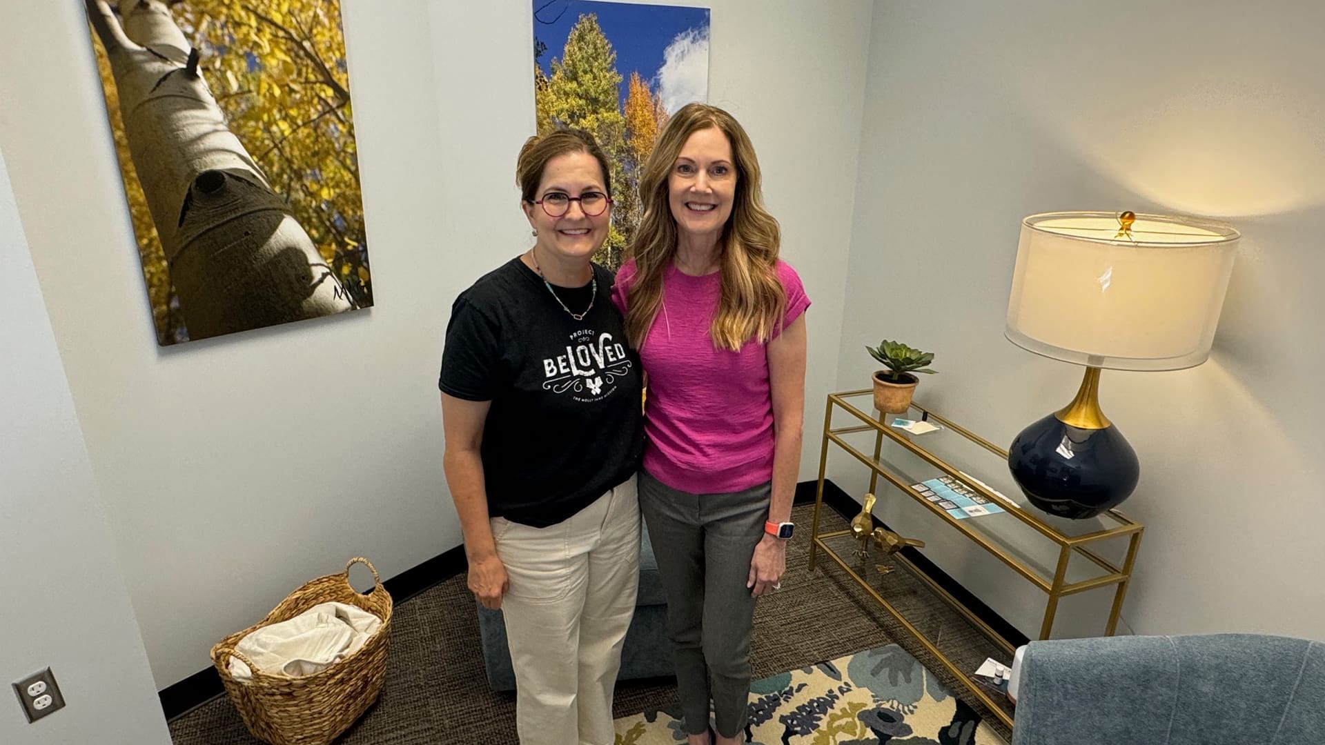 Two women stand side by side in a cozy room. One wears a black shirt, and the other wears a pink shirt. They are smiling. Behind them are two nature-themed paintings, and to the right is a lamp on a small table with a plant.