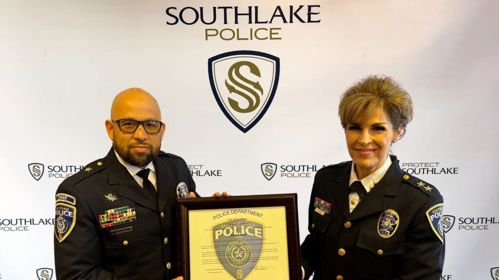 Two uniformed individuals stand in front of a backdrop with "Southlake Police" logos. They hold a framed certificate with a police badge design. Both wear decorated uniforms with insignias, standing side by side, facing the camera.