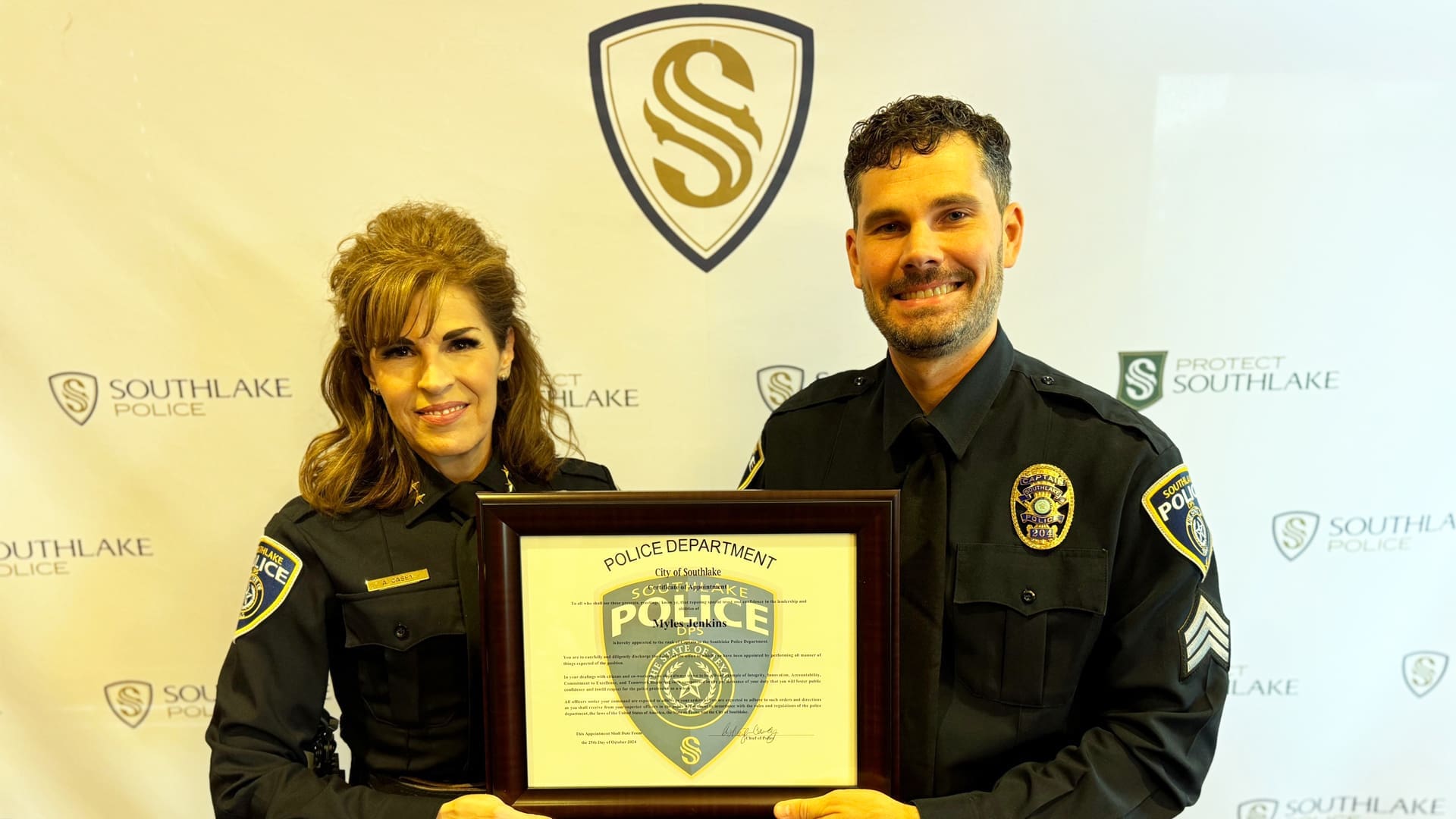 Two police officers smiling and holding a framed police department certificate. They are standing in front of a backdrop with the Southlake Police logo and text. Both are wearing police uniforms.