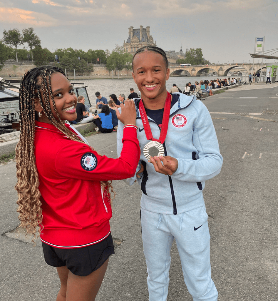 Two people smiling, one in a red jacket and the other in a light tracksuit with a medal. They stand near a river with people seated along the waterfront and a historic building in the background, under a cloudy sky.