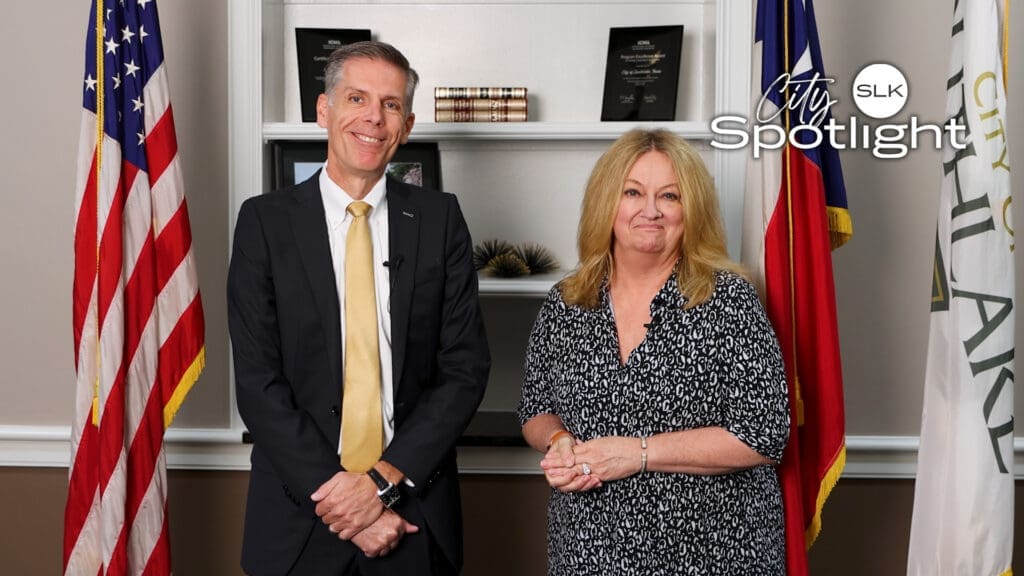 Two people are standing in an office with American and other flags behind them. They are smiling and dressed in formal attire. A "City Spotlight" logo is in the top right corner.