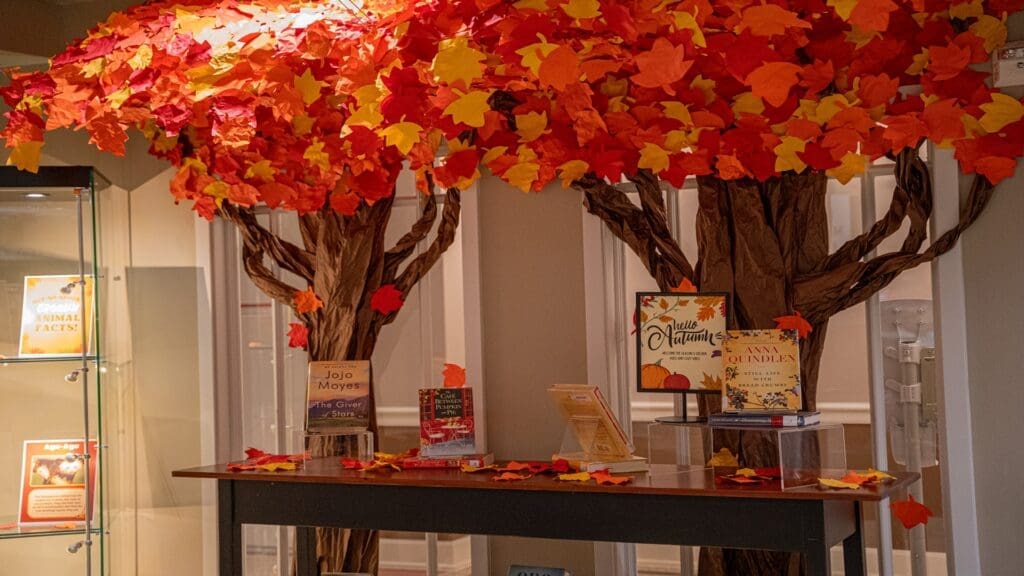 The image shows a paper-crafted fall tree with vibrant leaves. Below the tree, there are books displayed.