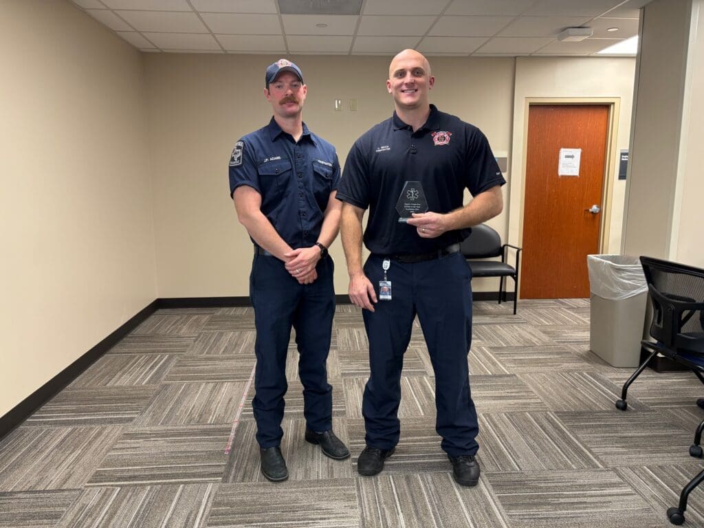 Two firefighters in uniform stand in a room with gray carpet. One holds a glass award. They are smiling, and the room has a closed wooden door and chairs.
