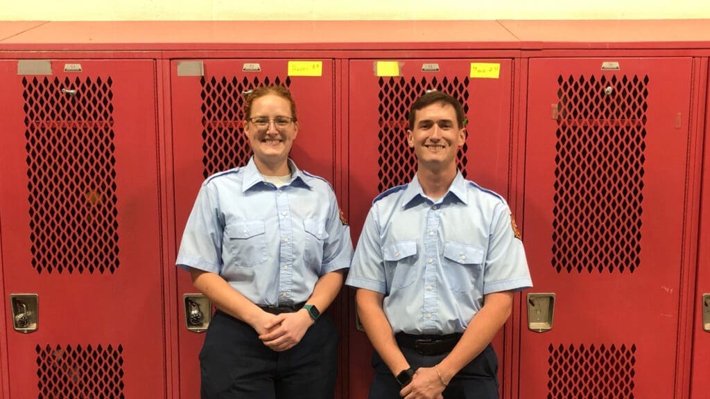 Two people in light blue uniforms stand in front of red lockers, smiling. They both have short sleeves and dark pants, with name tags visible on their shirts.