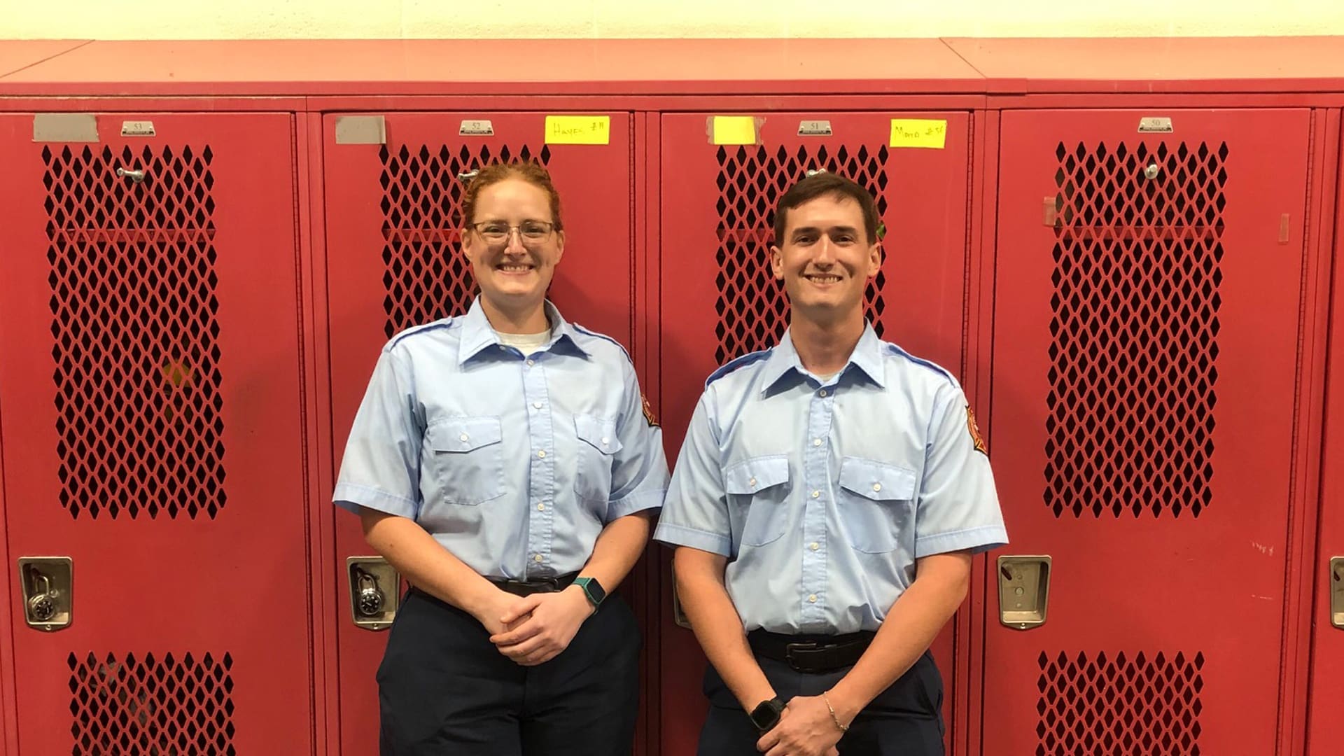 Two people in light blue uniforms stand in front of red lockers, smiling. They both have short sleeves and dark pants, with name tags visible on their shirts.