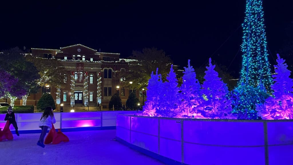 An illuminated ice rink at night featuring color-changing Christmas trees. A historic building adorned with lights is in the background. Two people appear to be skating or playing on the rink.