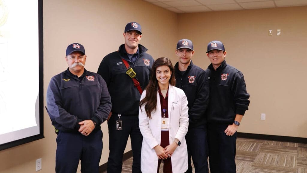 A woman in a white coat stands smiling with four people in dark blue uniforms and caps, who are also smiling. They are in a room with a carpeted floor and beige walls. A projection screen is partially visible to the left.