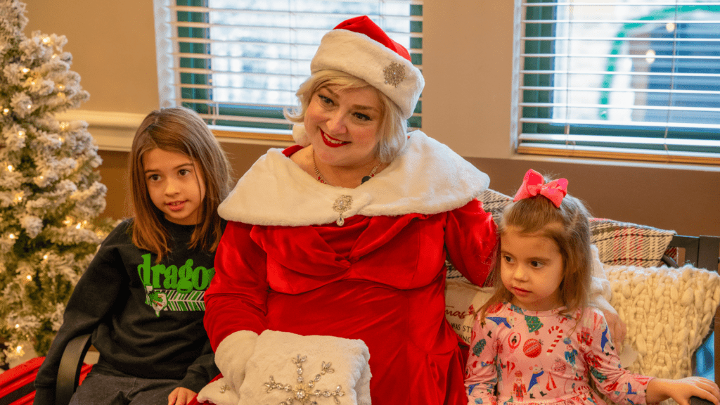 Mrs. Claus smiling with two young girls. All three are enjoying the moment, with festive decorations in the background