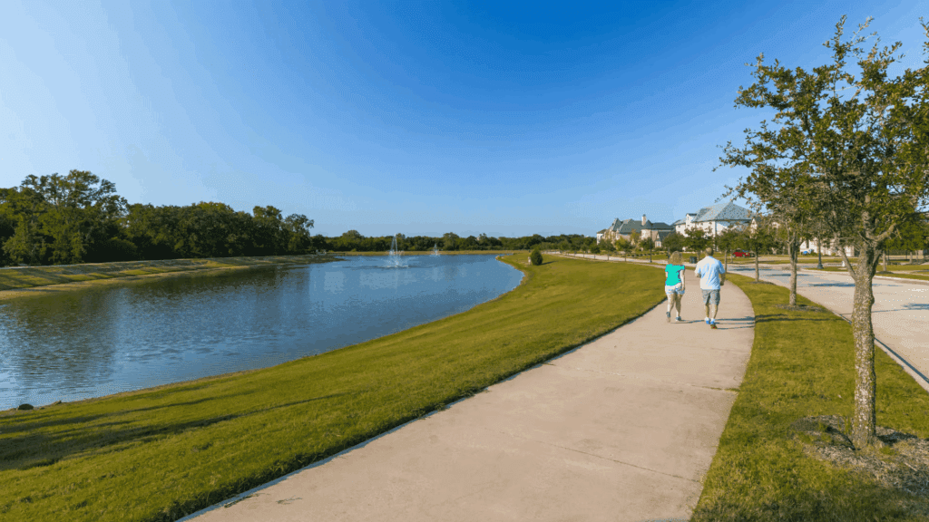 A couple walks along a paved path beside a calm pond with a fountain, surrounded by green grass, trees, and homes under a clear blue sky.