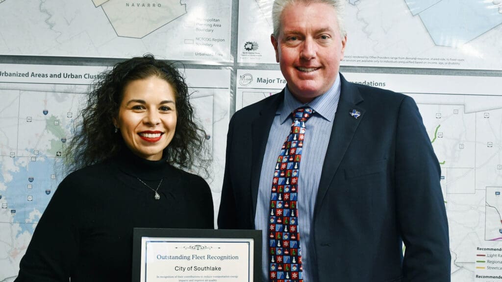 Two people stand smiling indoors in front of maps. The woman on the left holds a framed certificate that reads "Outstanding Fleet Recognition, City of Southlake." The man on the right is wearing a suit and patterned tie.