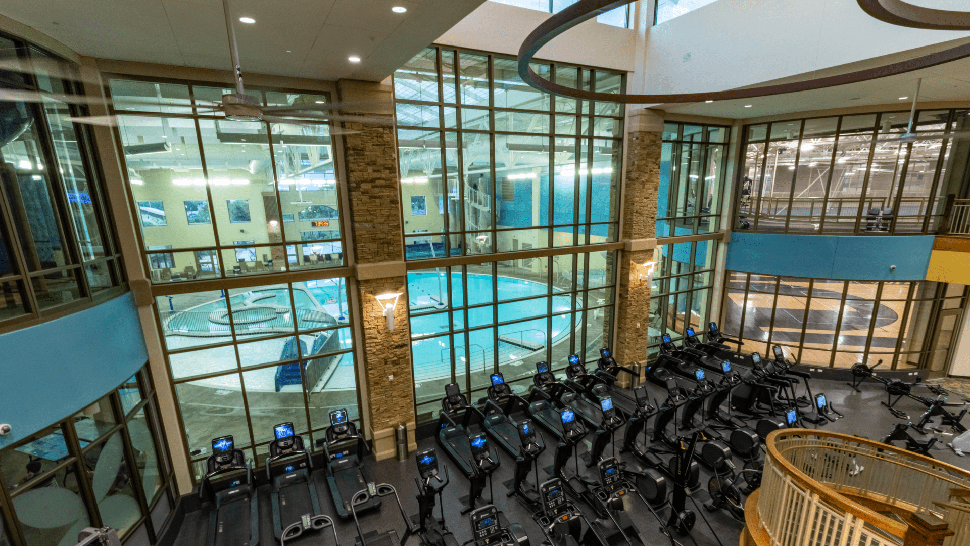 A spacious gym interior with a row of treadmills facing large windows. The windows overlook an indoor pool area with a turquoise circular pool. The space has high ceilings and modern architecture.