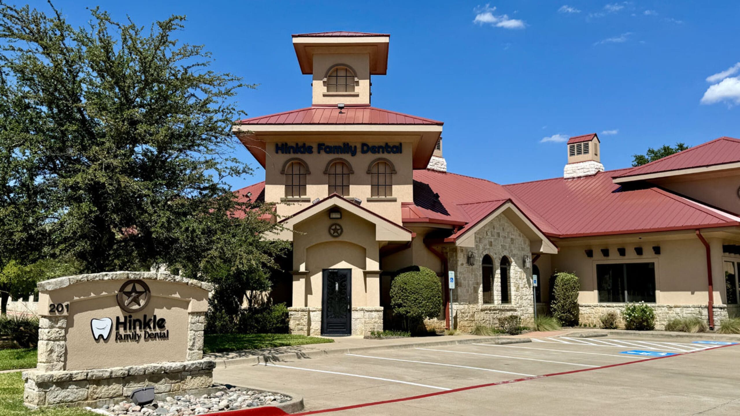 Image of a dental office building with a red roof and cream-colored walls. A sign in front reads "Hinkle Family Dental" and includes a tooth logo. Trees and shrubs surround the building, and there's a clear blue sky in the background.