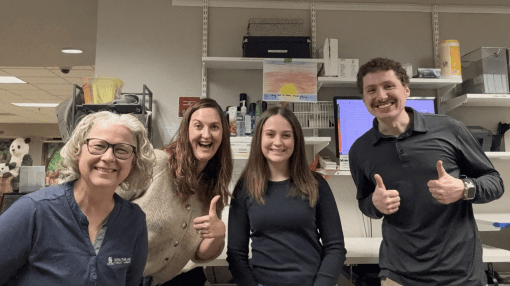 Four people smiling in an office setting. Two women on the left, one with glasses, and a woman and a man on the right giving thumbs up. Shelves with office supplies and a colorful drawing are behind them.