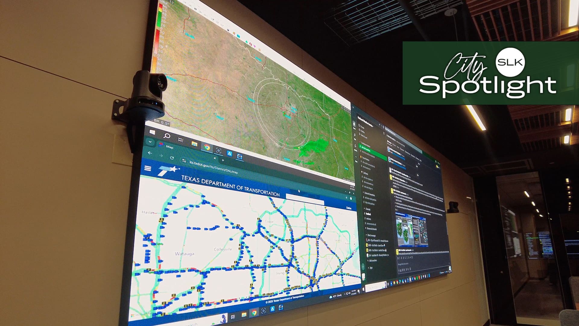A large digital display on a wall shows a satellite map and a traffic map from the Texas Department of Transportation. The screen is in a dimly lit room, with a "City Spotlight SLK" sign in the top right corner.