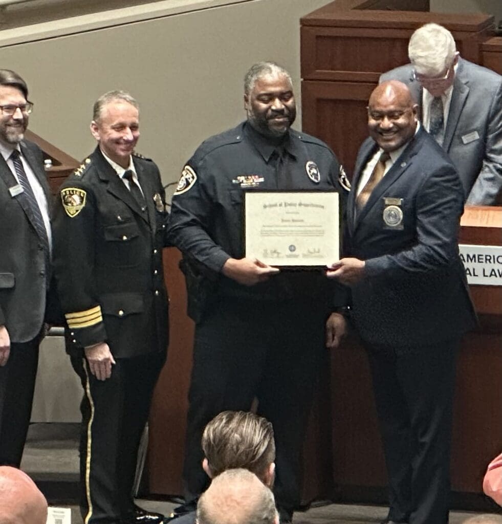 A group of five men stand indoors at a ceremony. One man in uniform holds a certificate with the help of another suited man. Two uniformed officers and one suited man stand nearby. A man in the background is seated at a podium.
