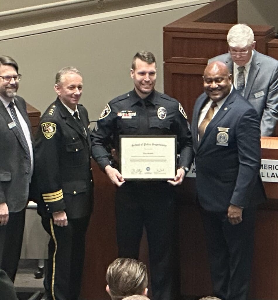 A group of five men stand together, with one man in a police uniform holding a certificate. They are all smiling, and a podium is visible in the background with a sign that partially reads "American Law.