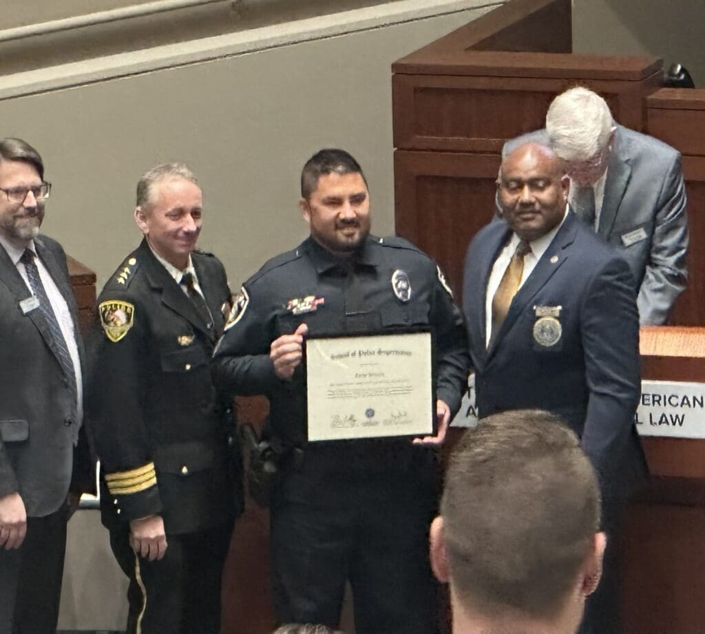 A group of five men are gathered. One, in a police uniform, holds a certificate. Another man in a suit stands to his right. A police officer stands to his left. Two other men in suits are behind a lectern. They appear to be at an awards or recognition ceremony.