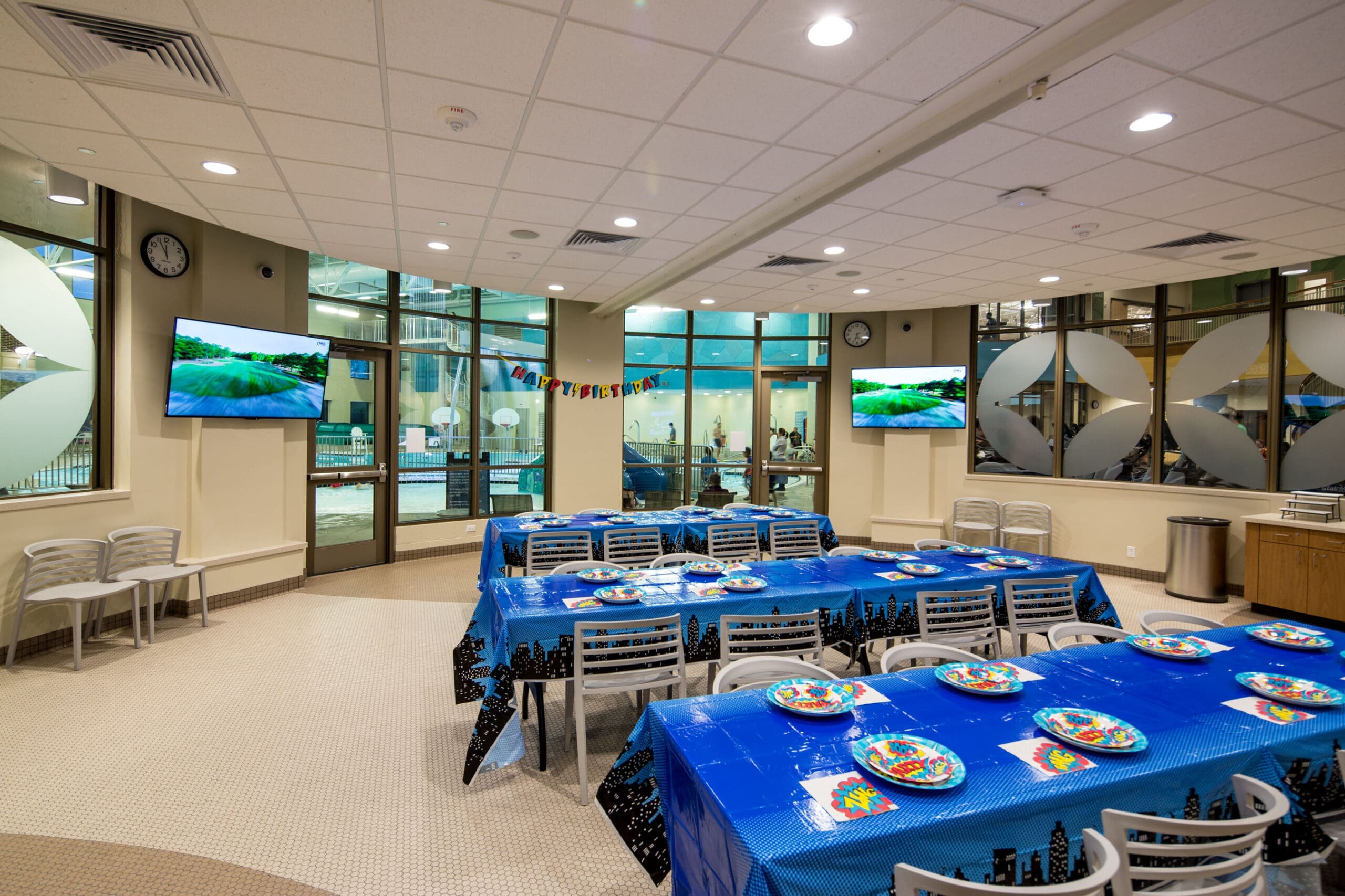 A decorated party room features tables draped in blue with colorful plates, ready for guests. "Happy Birthday" is spelled out in block letters. Windows reveal a pool area outside. Two wall-mounted TVs display a water scene. Gray chairs line the walls.