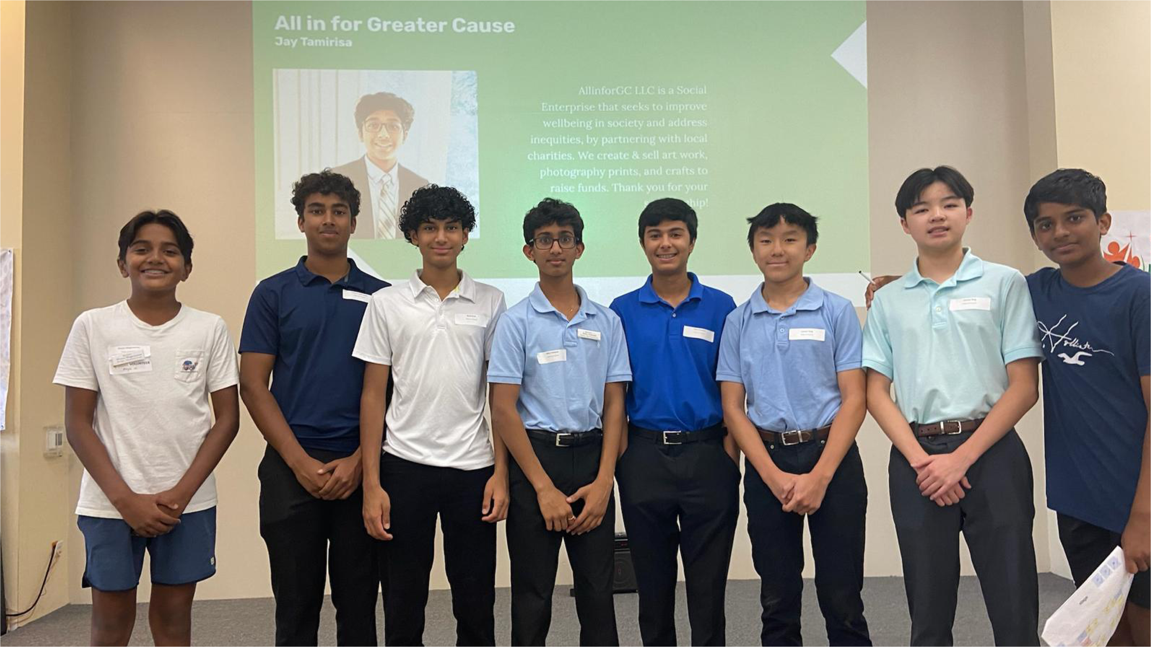 Eight young individuals stand in a group indoors, smiling at the camera. A presentation slide with text and an image of a person is projected behind them on the wall.
