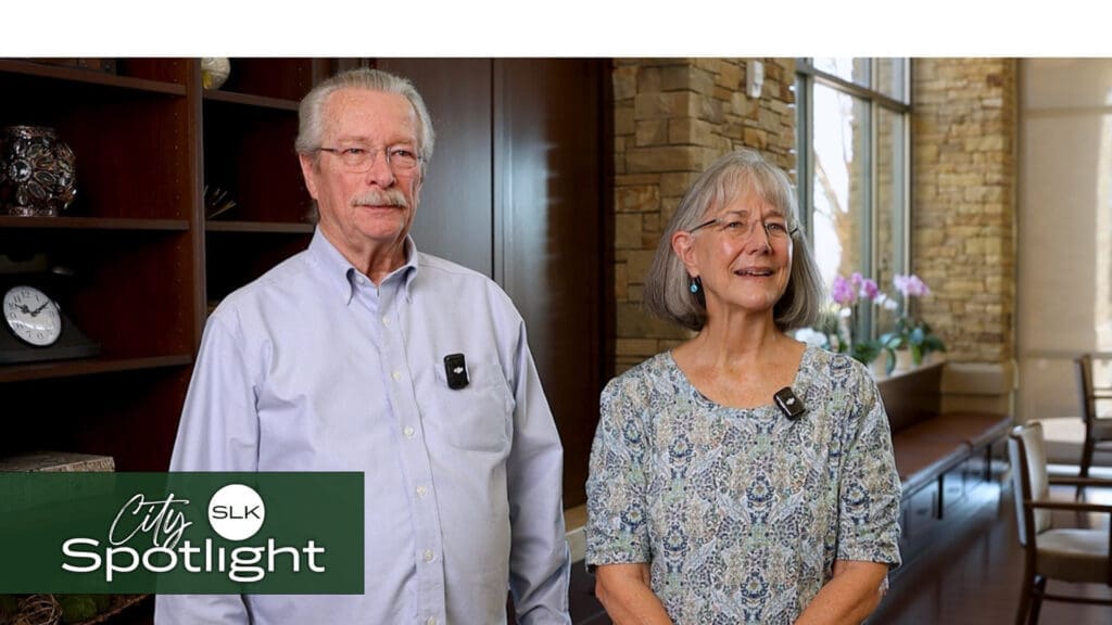 An older man and woman, both wearing light-colored clothing, stand indoors with microphones clipped to their shirts. A shelf with decorative items and a window showing plants are in the background. The image has a "City Spotlight" logo on the bottom left.