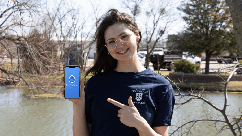 A person with long hair and glasses is smiling while holding and pointing to a smartphone displaying the EpicWater app. They are standing outdoors near a pond, wearing a navy blue t-shirt with "City of Southlake Public Works" written on it.