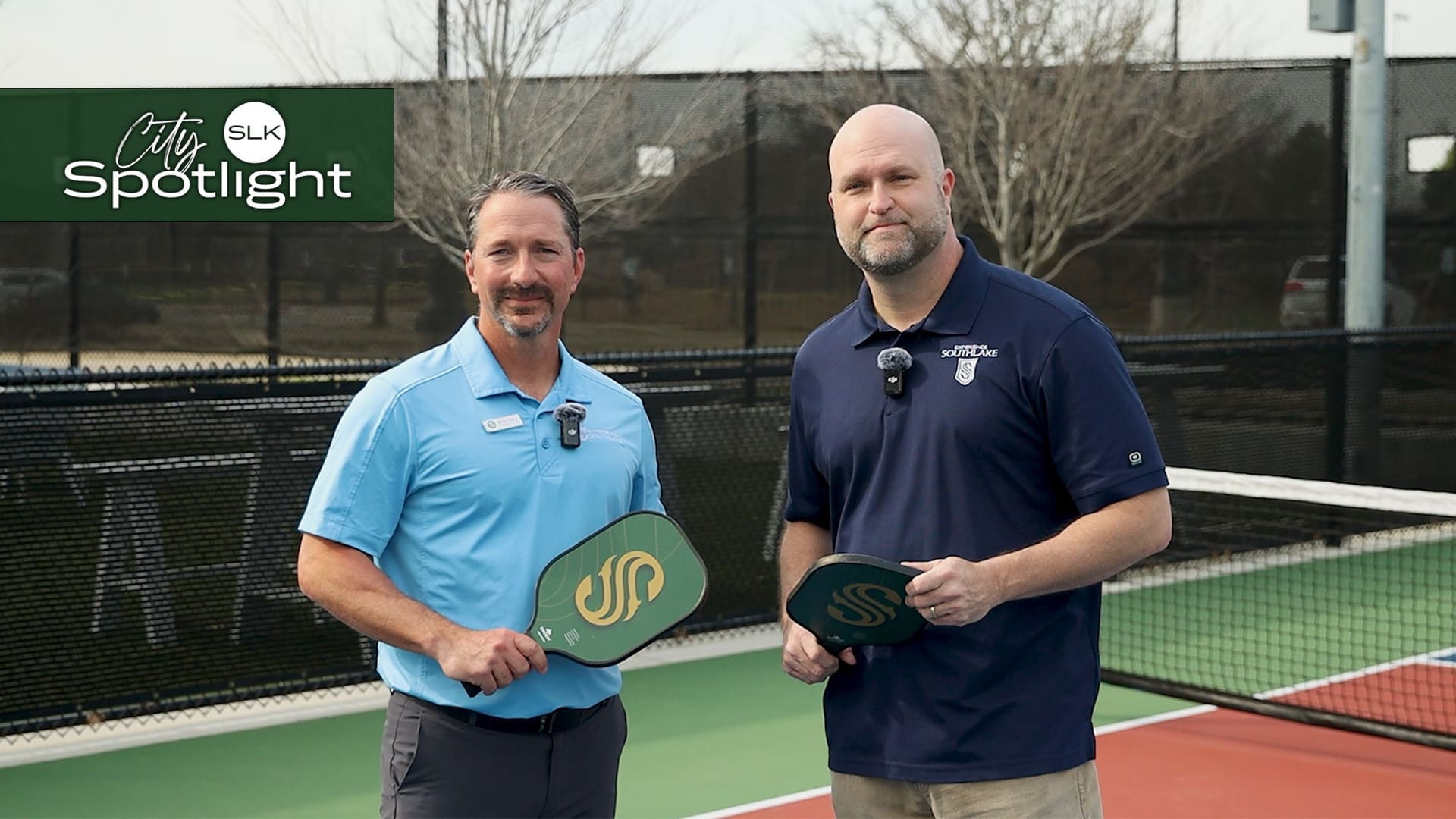 Pickleball Complex Manager Eric Clay and Mayor Pro Tem Randy Williamson standing on a pickleball court, each holding a paddle. They are dressed in casual attire with microphones resting on their shirts. The court has a net and trees are visible in the background. The image has a "SLK City Spotlight" logo in the corner.