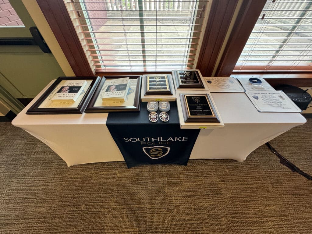 A table with a "Southlake Police" cover displays framed photos, awards, police badges, and certificates, arranged neatly. The backdrop includes large windows with blinds.