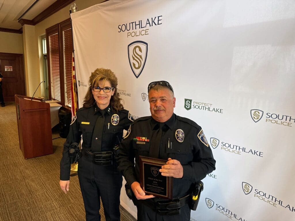Two uniformed police officers standing indoors. The officer on the right holds a plaque. A backdrop with "Southlake Police" and a logo is behind them. An American flag is partially visible on the left.