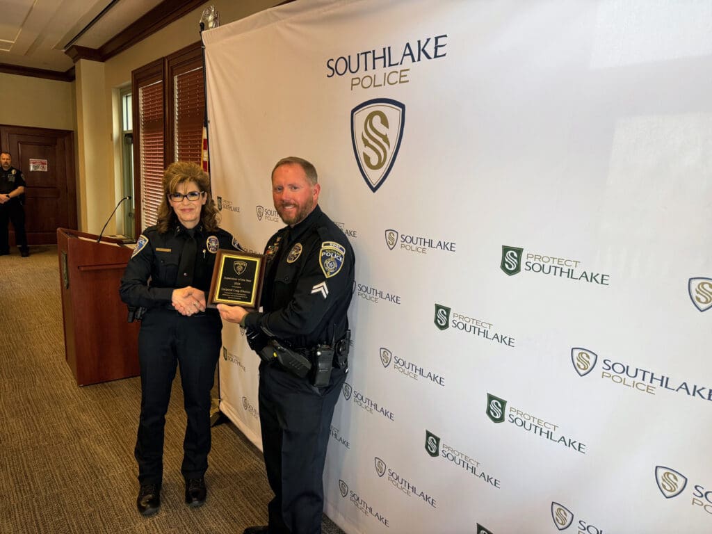 Two police officers in uniform stand indoors. One officer, holding a plaque, is being presented with an award by the other. A Southlake Police banner is in the background.