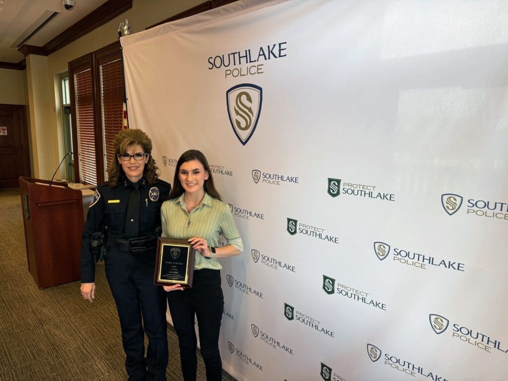 A police officer in uniform stands next to a woman holding a plaque. They are in front of a backdrop with the Southlake Police logo and text. The setting appears to be indoors, possibly at a police department event.