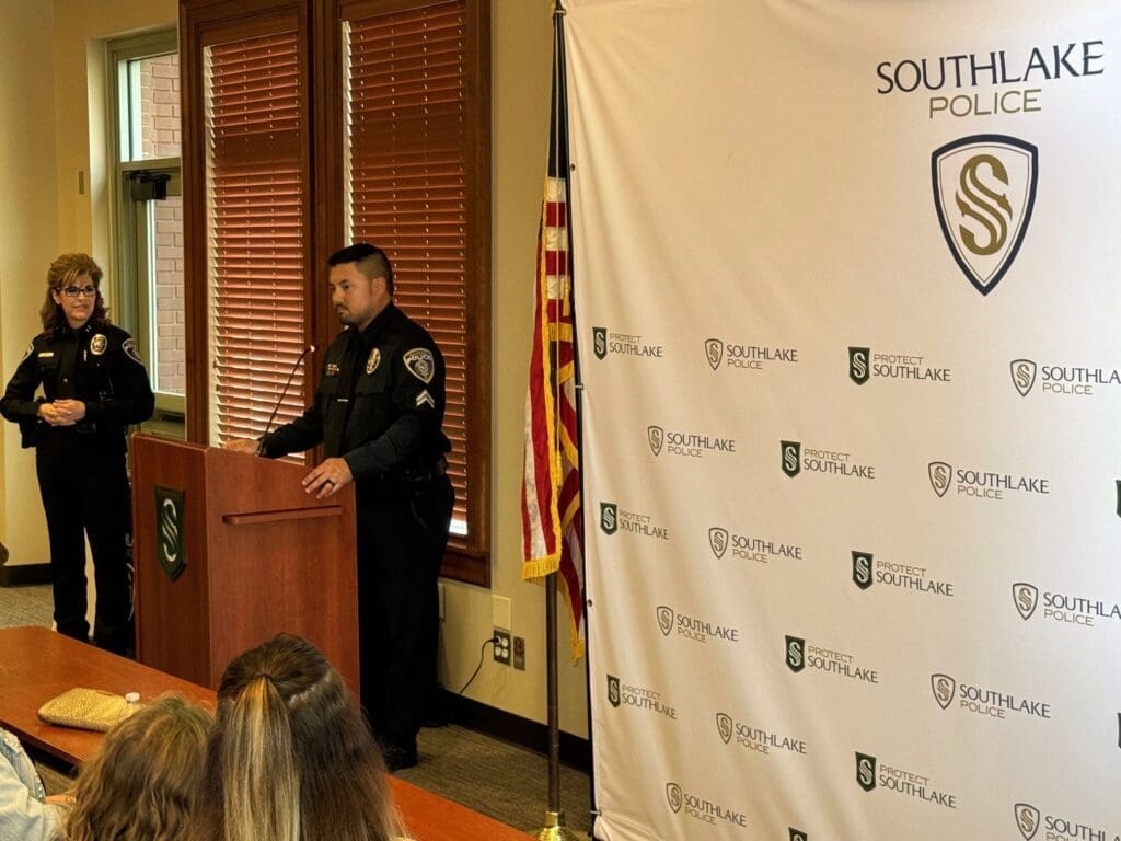 A police officer speaks at a podium with a "Southlake Police" logo. Another officer stands to the side. An American flag is nearby, and several people are seated in front of them. A banner with the police logo is in the background.