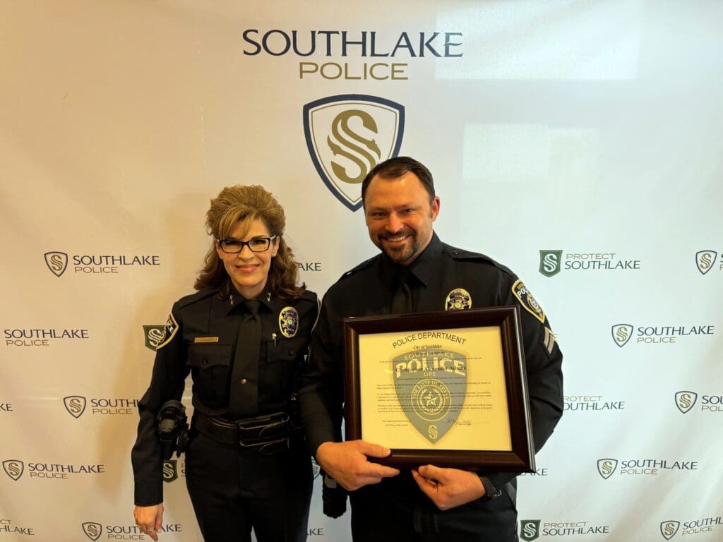 Two police officers stand in front of a Southlake Police backdrop. The male officer holds a framed certificate. Both are wearing dark uniforms with badges and are smiling at the camera.