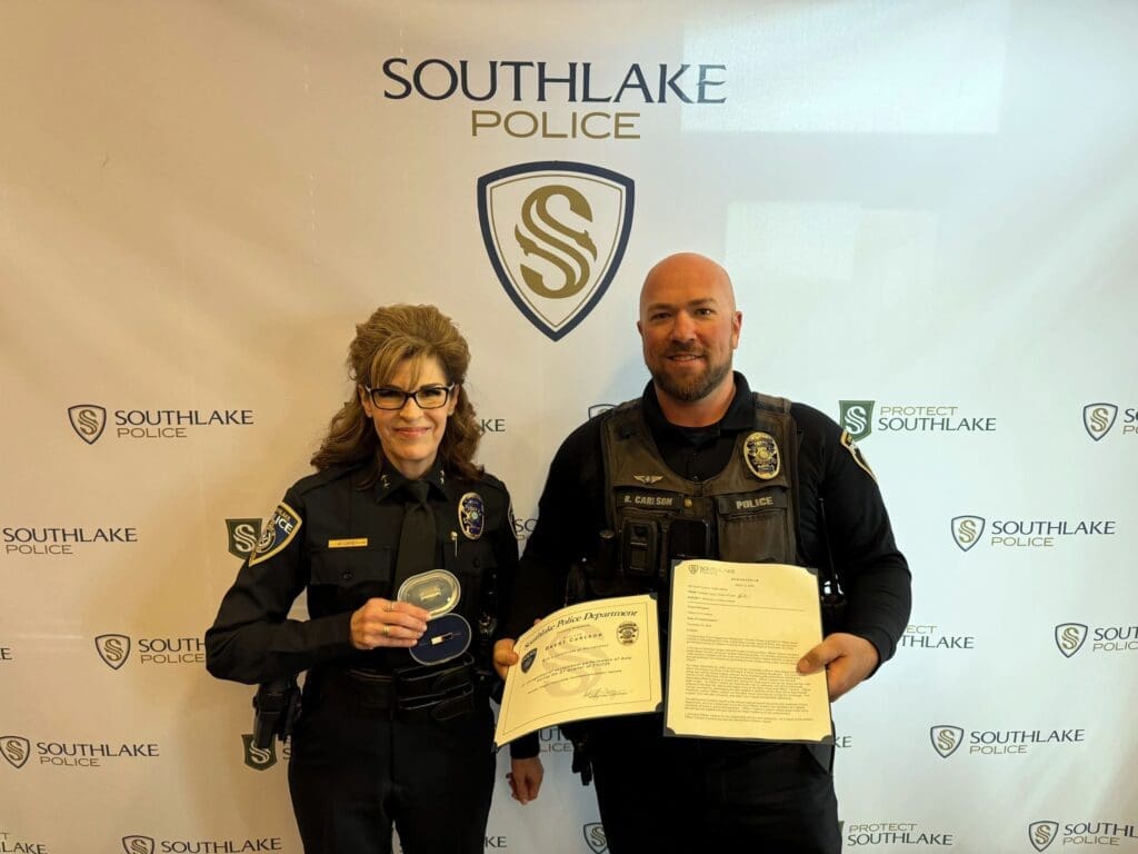 Two uniformed police officers stand smiling in front of a Southlake Police backdrop. The officer on the left holds a badge and a plaque, while the officer on the right holds a certificate. Both are wearing black police uniforms with badges.