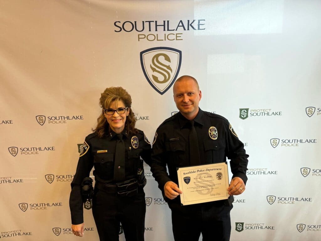 Two uniformed police officers stand in front of a Southlake Police backdrop. One officer is a woman on the left; the other is a man holding a certificate. Both are smiling.
