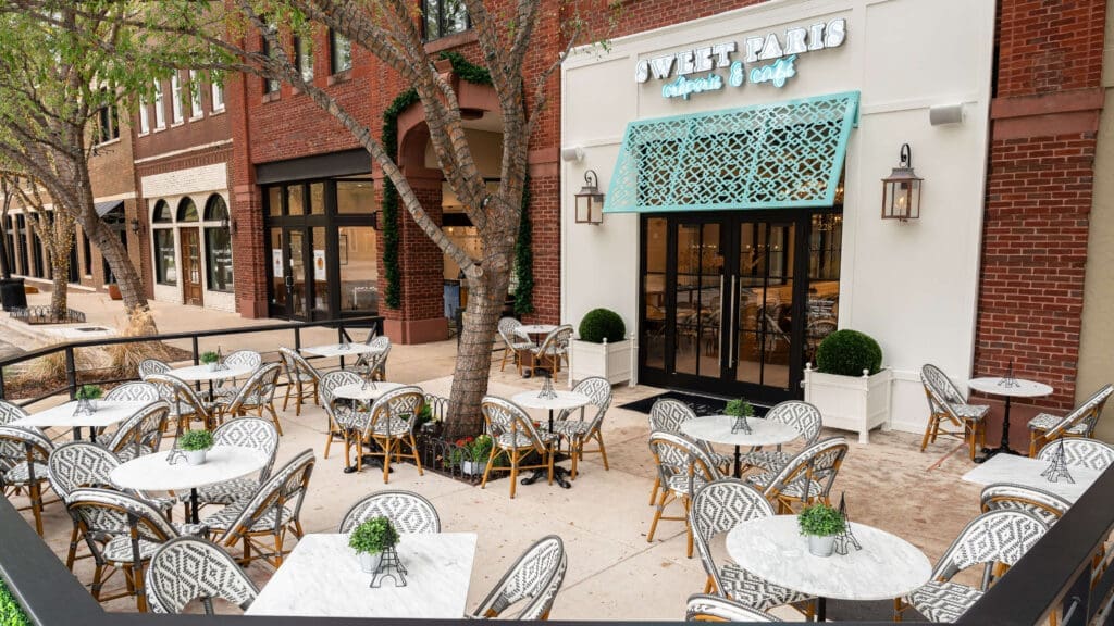 Outdoor seating area of Sweet Paris Crêperie & Café with white marble tables, patterned chairs, small potted plants on each table, and trees providing shade. The café has a teal sign and decorative awning.