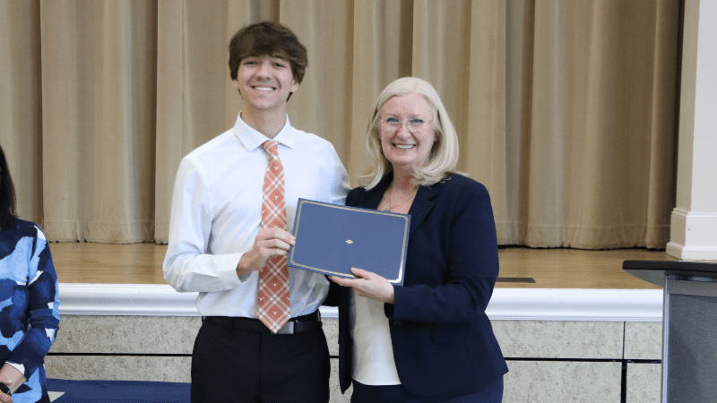 A young man in a white shirt and orange tie stands next to an older woman in a navy suit. Both are smiling and holding a certificate together, standing in front of beige curtains on a stage.