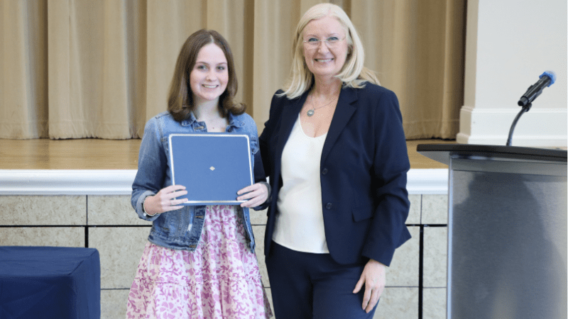 Two women stand indoors; one young woman in a pink dress holds a certificate and smiles. The other, an older woman in a navy suit, stands beside her, also smiling. A podium and microphone are visible on the right.