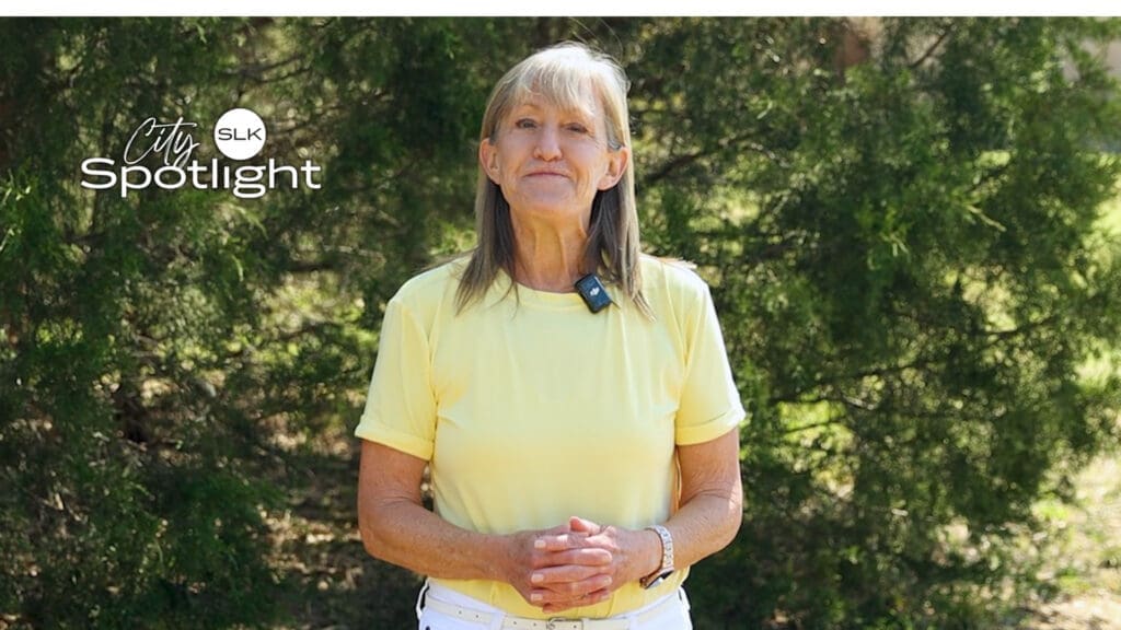 A person with long gray hair stands outdoors in front of greenery, wearing a yellow shirt. They have a microphone clipped to their shirt. The words "City Spotlight" appear in the upper left corner.