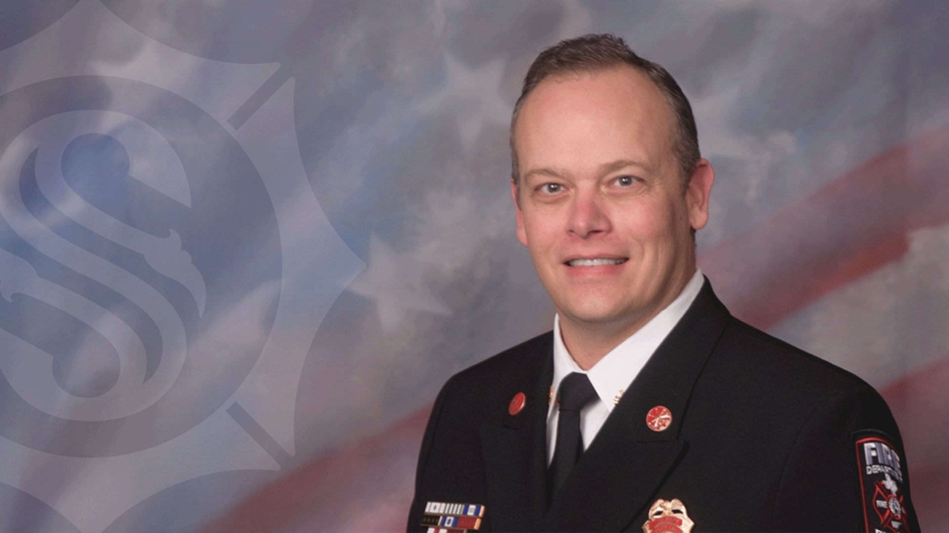 A firefighter in formal uniform, featuring various badges and pins, smiles at the camera. The background is a blurred patriotic theme with a subtle emblem on the left side.
