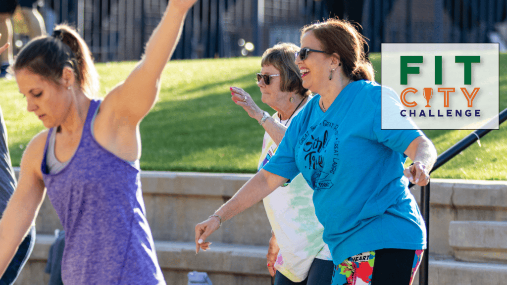 Three women exercising outdoors on a sunny day, smiling and moving energetically. In the corner, a logo reads "FIT CITY CHALLENGE" with a green and orange design. Grass and stairs are visible in the background.