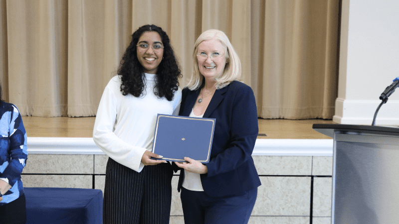 Two women stand together and smile at the camera; one is handing a certificate in a blue folder to the other in a brightly lit room with beige curtains in the background.