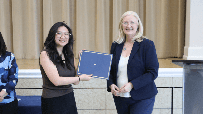 Two women stand indoors, smiling at the camera. The woman on the left holds a certificate, while the woman on the right, dressed in a navy suit, stands beside her. A podium is visible on the right.