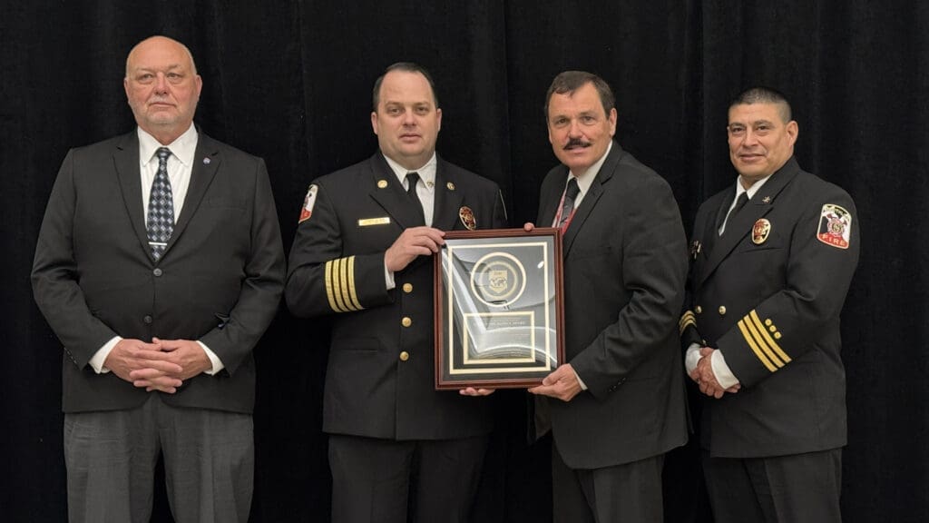 Four men in formal attire, including two in fire department uniforms, stand against a black background. One man holds a plaque with a badge emblem. They are all facing the camera.