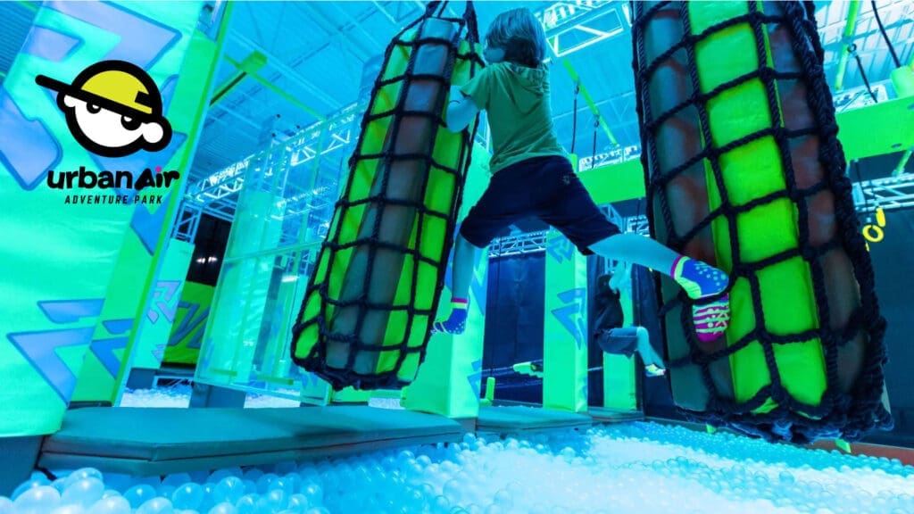 A child in athletic clothes climbs between two suspended nets above a pit filled with white plastic balls at an indoor adventure park. The scene is brightly lit with neon blue and green lights. Urban Air Adventure Park logo is visible.