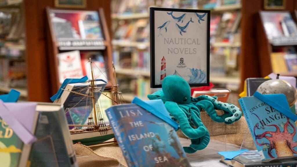 A bookstore display featuring nautical-themed novels. Books are arranged around a model ship and a plush octopus. A sign labeled "Nautical Novels" is visible in the center. The background shows more books and wooden shelves.