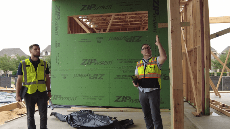 Two construction workers in safety vests inspect a partially built house, with green ZIP System sheathing visible. One person points upward while holding papers; the other stands nearby watching. Exposed wooden framing surrounds them.