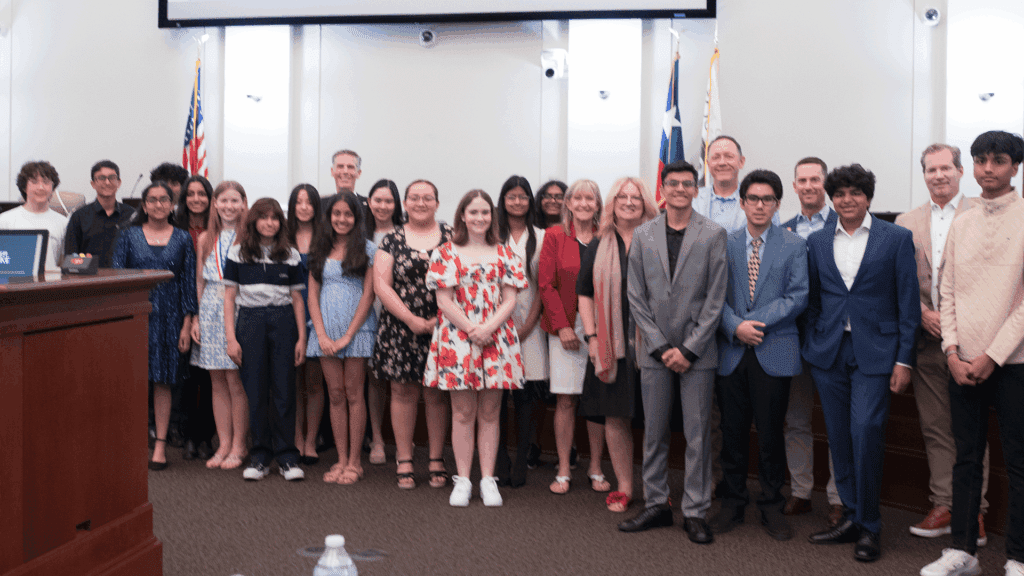 A group of students and adults stand together in a conference room, smiling at the camera. There are flags and a podium in the background, suggesting a formal or official event.