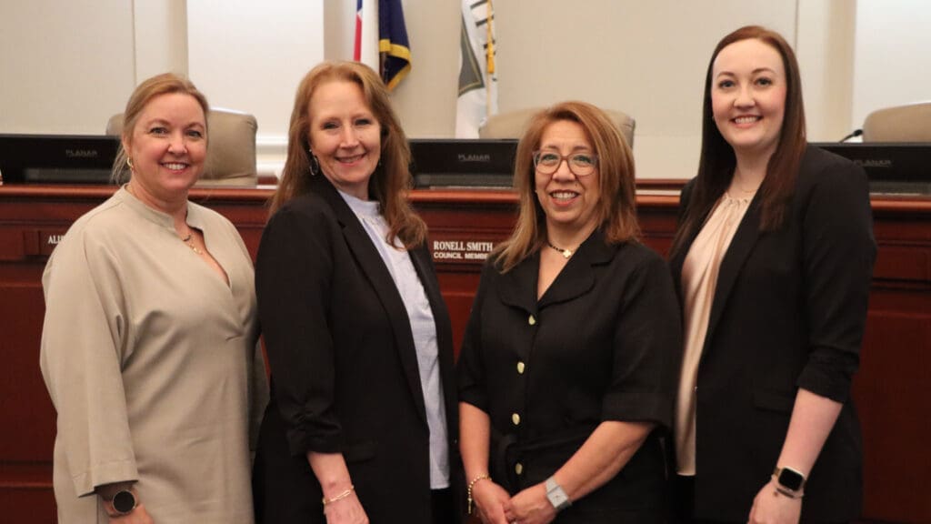 Four women stand side by side, smiling at the camera in a formal room with wooden desks and nameplates, suggesting a government or council setting.