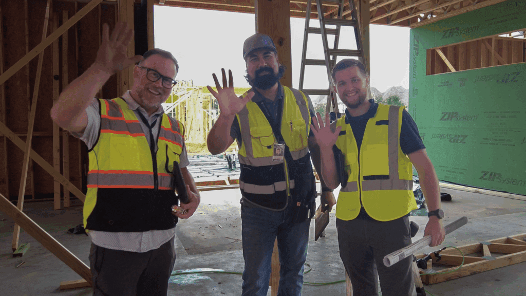 Three men wearing safety vests and holding blueprints smile and wave at the camera inside a building under construction, with wooden framing and a ladder visible in the background.