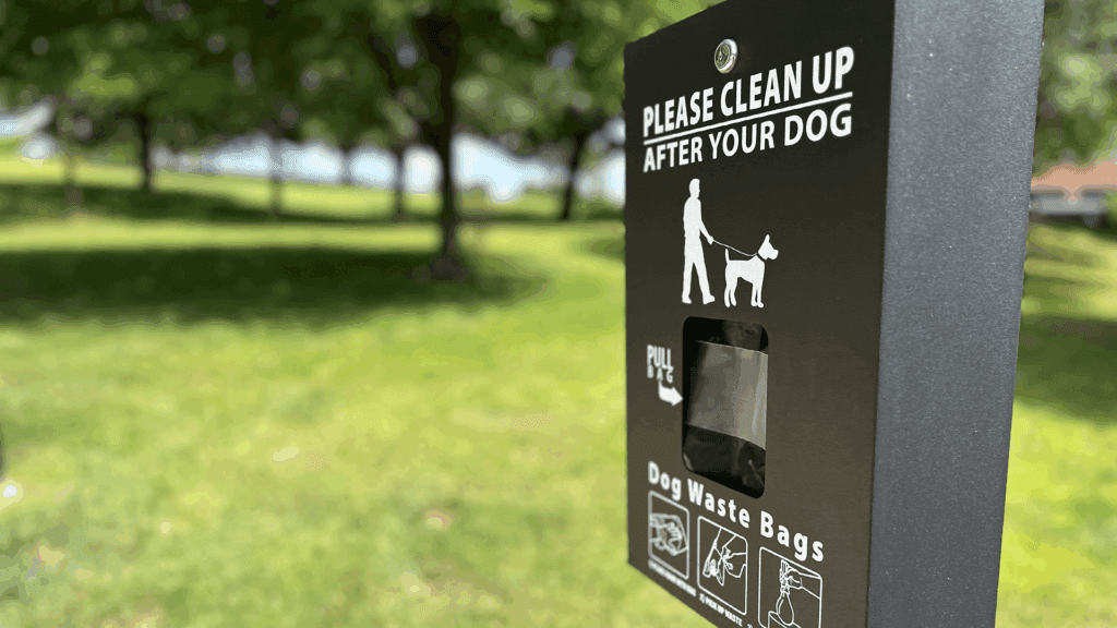 A black dispenser with "Please clean up after your dog" and "Dog waste bags" written on it is mounted in a grassy park area with trees in the background.